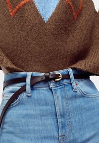 Suéter marrón de punto con patrones azules y naranjas, sobre unos pantalones de mezclilla azul claro, sujetos con un cinturón delgado de cuero negro y herrajes plateados.