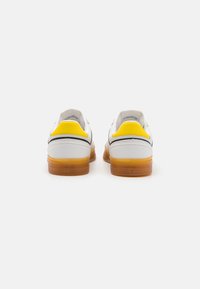 Zapatillas de cuero blanco con un acento en el talón amarillo, detalles en franjas negras y suela de goma. Acabado texturizado en el talón.