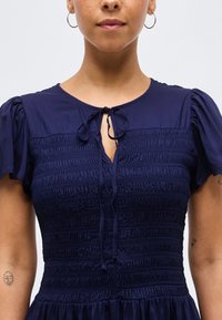 Γυναίκα που φοράει φόρεμα μπλε ναυτικό με σακουλωτό ύφασμα, κοντά φουσκωτά μανίκια, δεσίματα στον λαιμό και μικρά τατουάζ και στους δύο πήχεις.