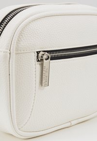 Borsa cosmetica in pelle bianca testurizzata con cerniera nera, caratterizzata da un'etichetta argento con la scritta "United Colors of Benetton" e un design curvo.