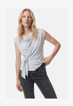 Blondynka nosząca jasnoszary bezrękawnik z drapowanym dekoltem i węzłem z przodu, zestawiony z czarnymi dżinsami.
