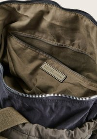 Bolsa de tela negra con un interior suave y texturizado. Presenta un bolsillo con cremallera y una etiqueta que dice "DENIM TOM TAILOR". Forrada en verde oliva.