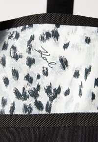 Svart tote-veske med en teksturert stropp. Innvendig har den et hvitt stoff med svarte penselstrøk leopardprikker. Inkluderer en signatur i kursiv.