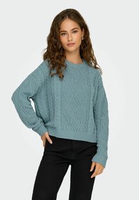 Pull en maille bleu clair avec un motif de losanges texturé, poignets et ourlet côtelés, associé à un pantalon noir. Coupe décontractée, manches longues.