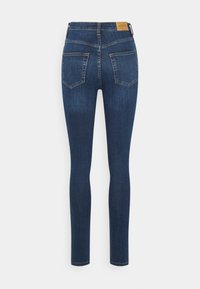 Jeans skinnies de tiro alto en azul oscuro con dos bolsillos traseros y un parche de cuero marrón en la cinturilla sobre un fondo blanco.