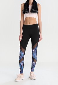 Černé legíny s modrým tropickým vzorem a síťovými detaily; v kombinaci s růžovým cropped topem a světle růžovými teniskami. Ručník na ramenou.