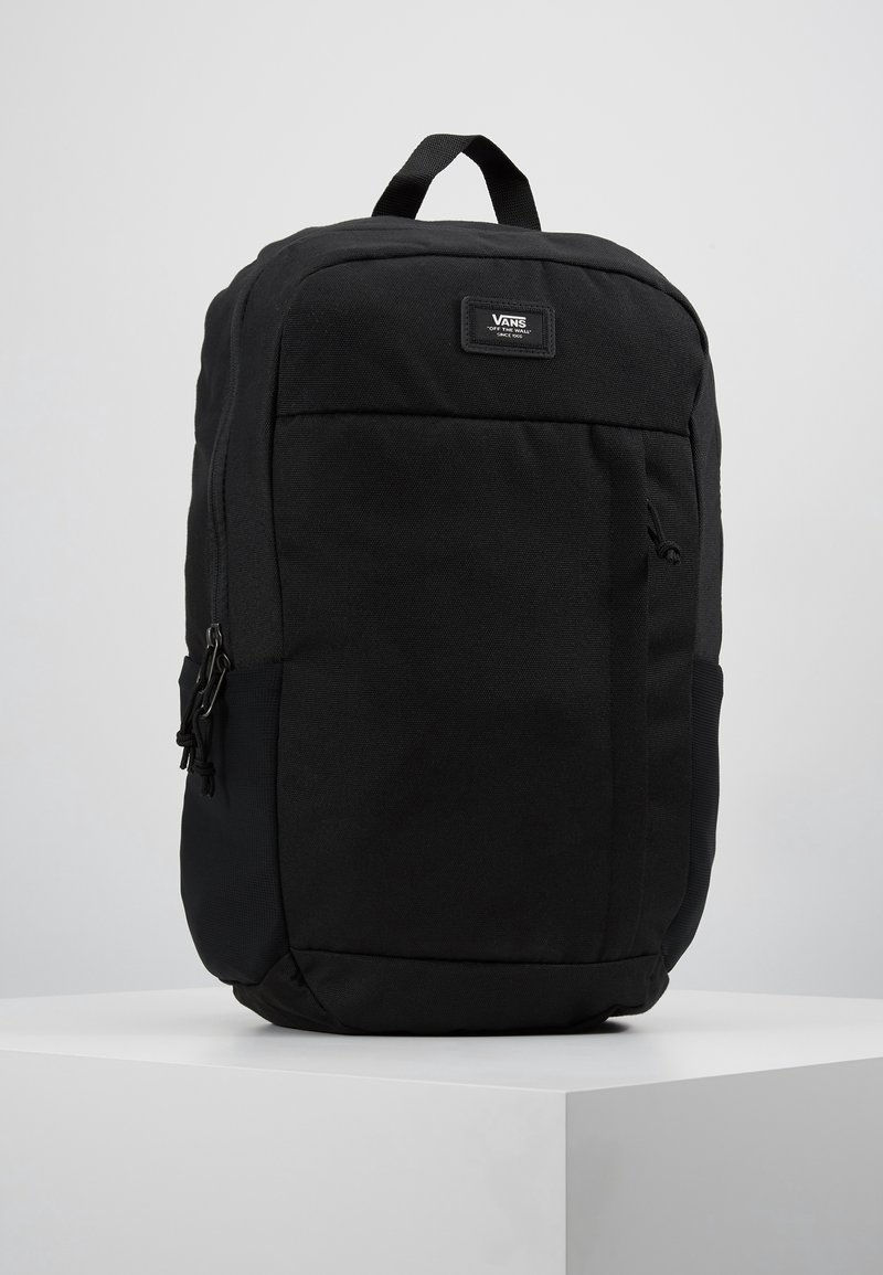 Sac à dos noir Vans avec une poignée supérieure, une poche avant zippée et un écusson avec le logo Vans sur une surface blanche unie.