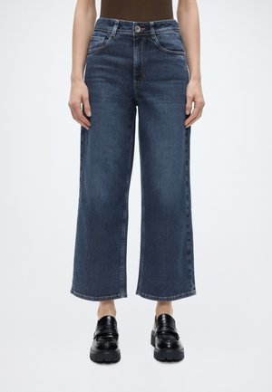 Kobieta ubrana w ciemnoniebieskie, szerokie, skrócone dżinsy z przednimi kieszeniami oraz czarne loafersy na platformie, stojąca na jednolitym tle.