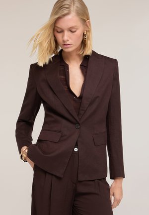 Vrouw met blond haar die een donkerbruine op maat gemaakte blazer en bijpassende broek draagt, geaccentueerd met gouden oorbellen en armbanden, kijkt naar beneden.