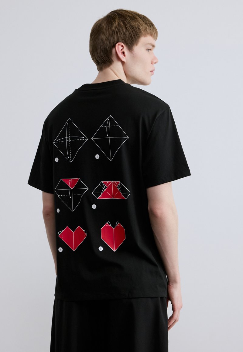 Μαύρο βαμβακερό μπλουζάκι με λευκές και κόκκινες εικονογραφήσεις οριγκάμι στην πλάτη, που περιλαμβάνουν γεωμετρικά σχήματα και καρδιές, με άνετη εφαρμογή.
