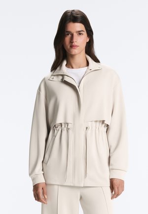 Femeie cu păr lung și întunecat, purtând o jachetă bej deschis cu fermoar și șnur în talie și pantaloni asortați, stând pe un fundal simplu.