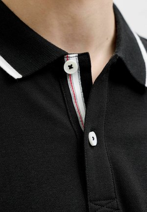 Fekete galléros póló közeli képe, fehér gombokkal és piros csíkkal díszített fehér szegéllyel a gombsor belsejében.