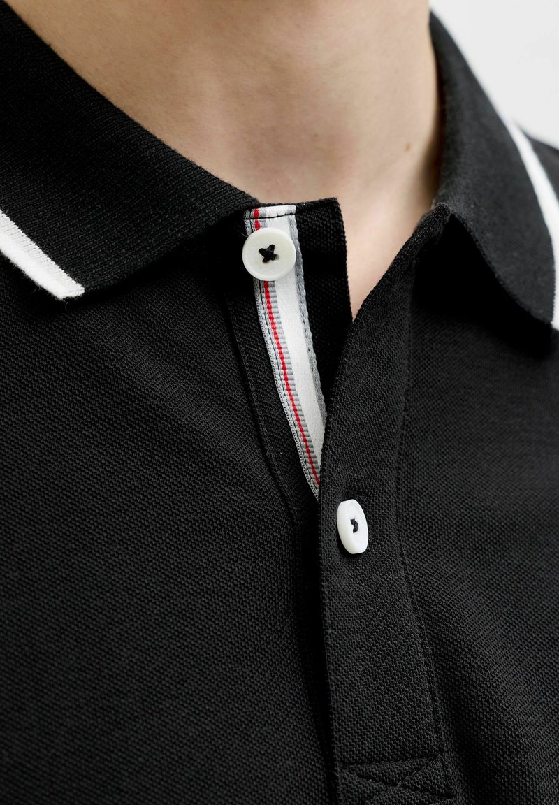 Prim-plan al gulerului unui tricou polo negru cu nasturi albi și tiv interior cu dungi albe și roșii pe fanta nasturilor.