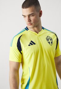 Żółta koszulka sportowa z granatowymi akcentami, z dekoltem w serek i krótkimi rękawami. Zawiera teksturowany wzór oraz emblemat Szwecji na klatce piersiowej.