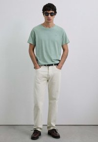 Camiseta de algodón verde claro con mangas cortas, combinada con pantalones color crema y zapatos de cordones marrón oscuro. Cinturón negro y gafas de sol incluidas.