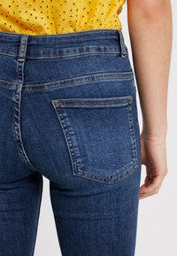 Mörkblå denimjeans har en åtsittande design med bakfickor och synlig sömnad. Texturen verkar slät med lätta blekningar.