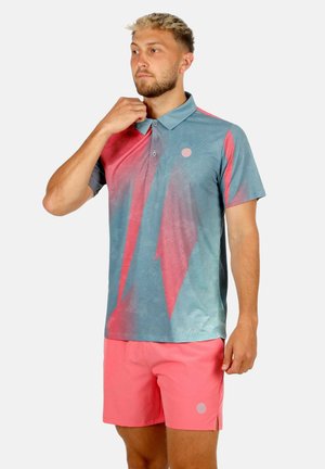Hombre con barba ajustándose el cuello, vistiendo una camisa polo de manga corta con degradado azul y rosa y pantalones cortos rosa coral sobre fondo blanco.