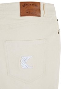Lähellä vaaleanbeige housun tasku, jossa on valkoisella brodeerattu "K"-logo ja ruskea nahkapatche, jossa lukee "Karl Kani" vyötäröllä.