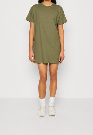 Femme portant une robe T-shirt oversize vert olive associée à des chaussettes blanches mi-mollet et des baskets blanches, debout devant un fond uni.