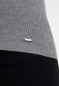 Harmaa ribbineulottu villapaita, jossa on hopeiset metallitarra "Morgan"-merkillä helmassa. Tekstuuri on pehmeä, ja se kontrastoi alla olevan sileän mustan kankaan kanssa.