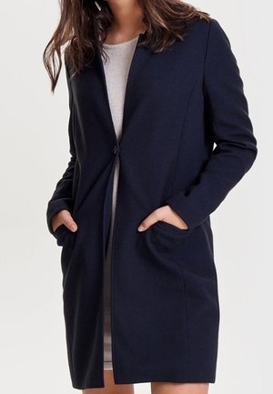 Frau trägt einen knielangen, dunkelblauen Mantel mit einem Knopf und hat die Hände in den Taschen, über einem hellgrauen Kleid.