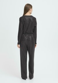 Czarny kombinezon z cekinami, z długimi rękawami, o luźnym kroju, z wzorem w jodełkę i elastycznym paskiem w talii, pokazany od tyłu na jasnym tle.