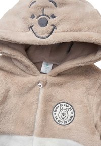 Felpa per neonati di colore beige, con cappuccio peloso e un volto di orsetto sorridente sul cappuccio. Sul petto è presente un patch circolare con il volto di un orsetto e del testo.