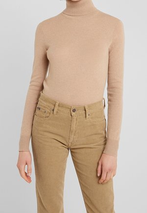 Person trägt einen beigen Rollkragenpullover, der in eine hoch taillierte, hellbraune Cordhose gesteckt ist, mit entspannt an den Seiten hängenden Händen.