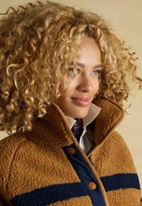 Brūns šerlinga mētelis ar tumši zilu svītru, augstu apkakli un brūnu apdari. Tajā ir divi lieli pogas, tas valkāts uz apkakles krekliņa.