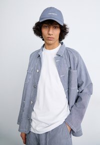 Striped blue and white jacket with large buttons, white t-shirt, and gray cap featuring logo. Casual fit and lightweight fabric.
