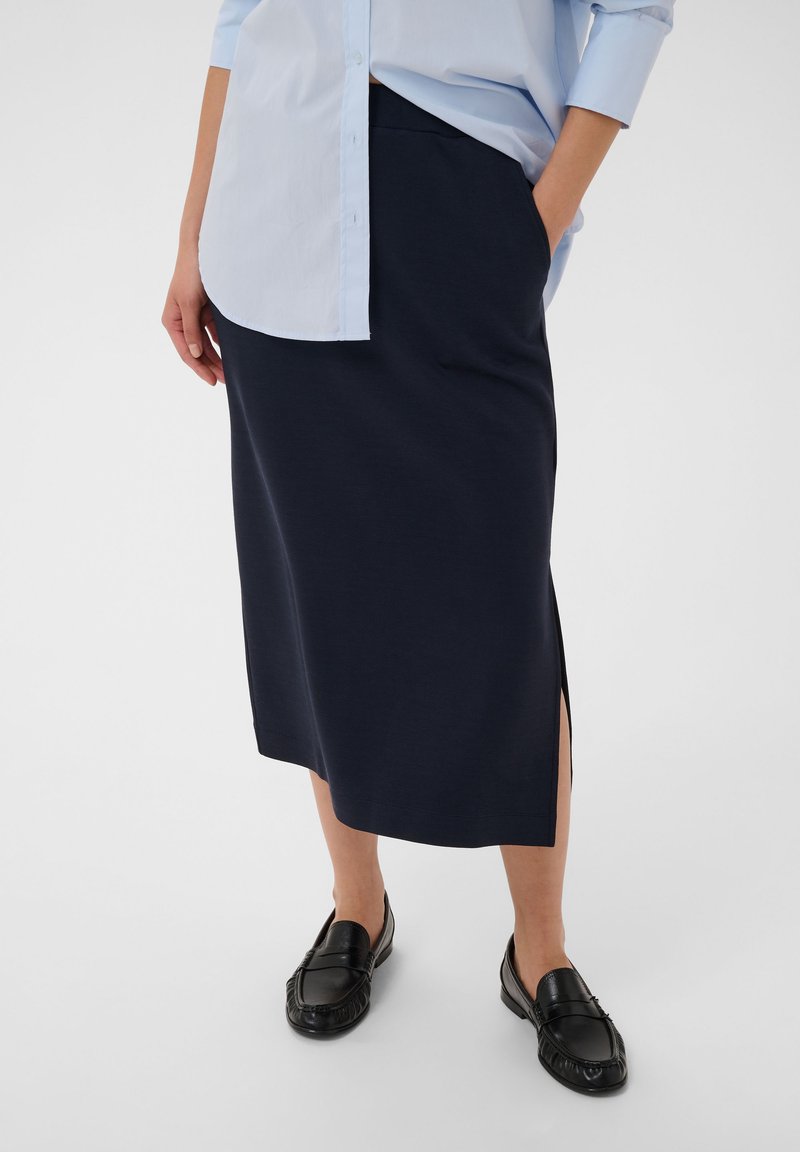Femme portant une jupe midi bleu marine fendue sur le côté, une chemise bleu clair boutonnée et des mocassins en cuir noirs, debout avec une main dans la poche.