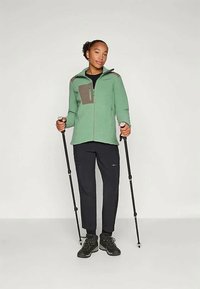 Πράσινο φλις μπουφάν με καφέ λεπτομέρειες, τσέπη με φερμουάρ, συνδυασμένο με μαύρο παντελόνι και μπότες. Άτομο κρατάει μπατόν πεζοπορίας.