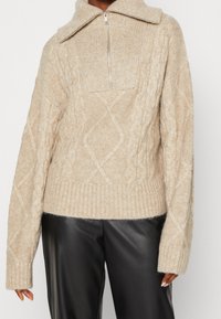 Beiger Strickpullover mit geripptem Kragen und halbem Reißverschluss, mit Zopfmuster und entspannter Passform. Kombiniert mit schwarzen Lederhosen.