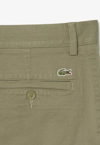 Gros plan sur la poche d'un pantalon kaki avec un bouton et un petit logo de crocodile vert brodé au-dessus de la poche.