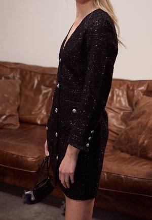 Frau trägt ein schwarzes, glitzerndes, knöpfe versehenes Kleid und hält eine kleine schwarze Handtasche, steht neben einem braunen Ledersofa.