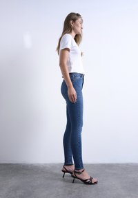 Γυναίκα που στέκεται πλαγίως, φοράει λευκό t-shirt, μπλε στενά τζιν, μαύρα πέδιλα με τακούνι, μπροστά σε απλό ανοιχτό φόντο.