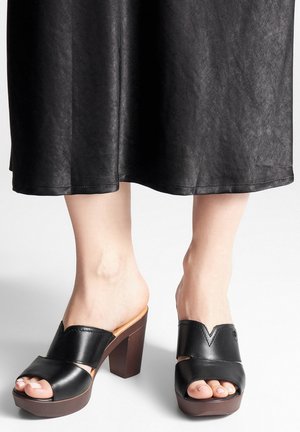 Mule cu toc din piele neagră, cu vârf deschis, design în formă de V și toc din lemn. Textură netedă și accenturi de cusături vizibile.
