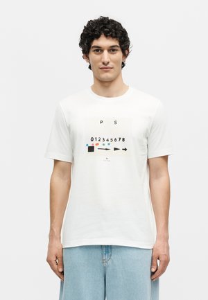 Νέος άνδρας με σγουρά μαλλιά που φοράει λευκό T-shirt με αφηρημένο σχέδιο που περιλαμβάνει νούμερα, βέλη και πολύχρωμες κουκκίδες, σε συνδυασμό με ανοιχτό μπλε τζιν.