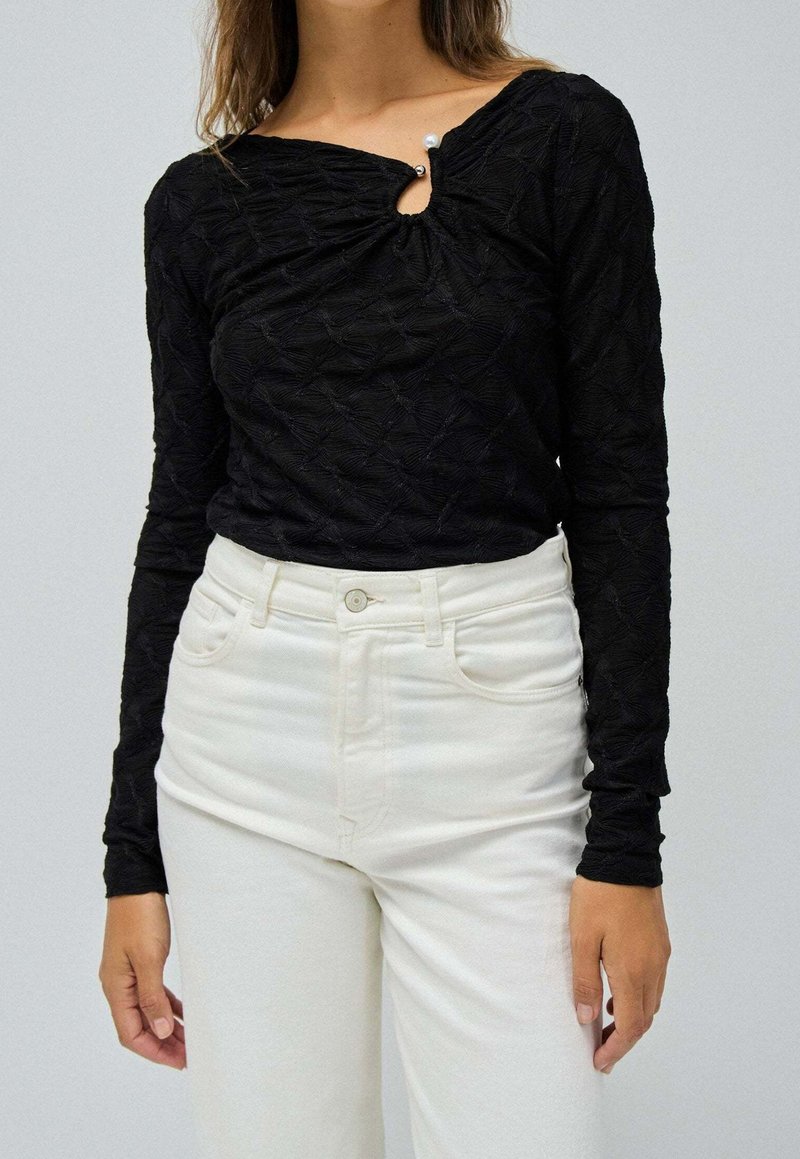 Femme portant un haut texturé noir à manches longues avec une petite ouverture et un pantalon blanc, debout devant un fond clair uni.