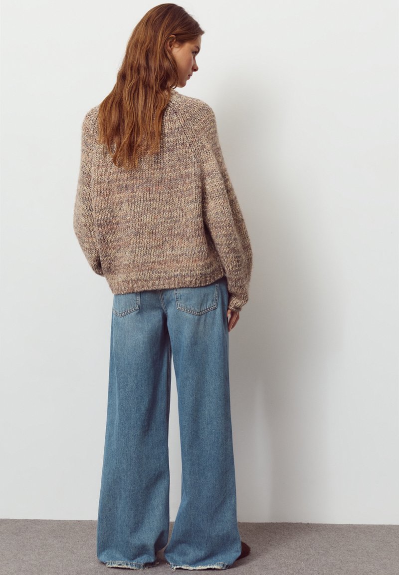 Ylimitoitettu beige neule, jossa on struktuurinen neulos, ja leveät siniset farkut, joissa on rispaantunut helma, esillä mallin selkäpuolelta.