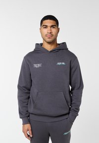 Uomo con felpa con cappuccio grigio scuro e pantaloni della tuta coordinati con piccoli loghi di testo bianchi e blu su petto e coscia, in piedi su sfondo bianco.