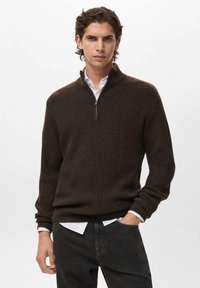 Maglione marrone a coste con colletto zip, realizzato in tessuto testurizzato. Indossato sopra una camicia bianca, con jeans scuri e polsini ben visibili.
