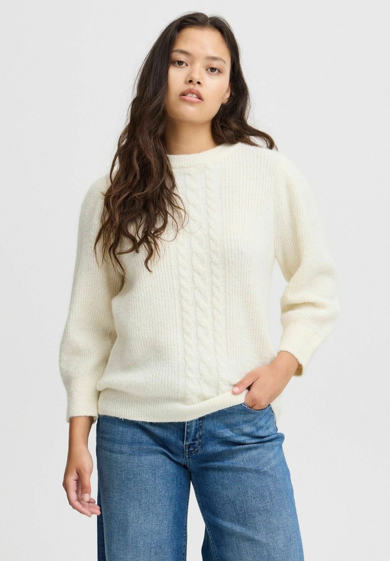 Pull en tricot torsadé de couleur crème, avec un col rond et des manches 3/4. Texture douce avec des accents côtelés. Associé à un jean bleu.