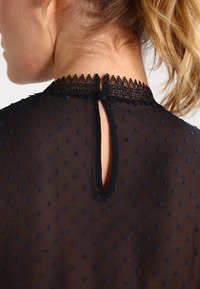 Schwarze Bluse mit Pünktchenmuster, Rückenteil aus strukturiertem Spitzenkragen, Schlüsselöffnungsdetail und durchsichtigen Stoff. Der Ausschnitt ist mit einer feinen Spitzenkante verziert.