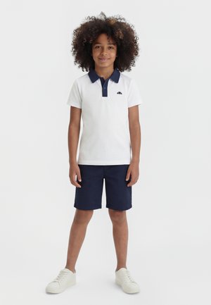 Barn med krøllete hår iført en hvit polo med marineblå krage, marineblå shorts og hvite sneakers, stående foran en ensfarget hvit bakgrunn.