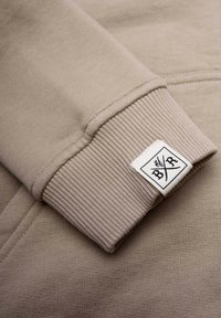 Ljusbeige sweatshirtärm med ribbat mönster, försedd med en broderad logotags i svart på vit bakgrund. Slät tygfinish.