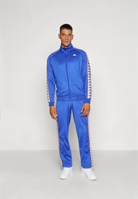 Kék tréningruha sima, könnyű anyagból. Kontrasztos csíkkal és logóval a dzsekiben és a nadrágon. Klasszikus szabású, cipzáras záródással.