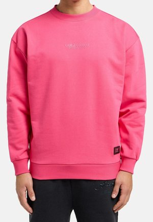 Person trägt ein leuchtend pinkes Sweatshirt mit langen Ärmeln und dem Schriftzug "Carlo Colucci Milano 1976" mittig auf der Brust sowie schwarze Hose.