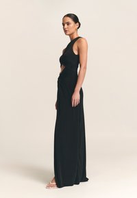 Musta iltapuku, jossa on kiertynyt leikkaus vyötäröllä, hihaton malli, lattiaan ulottuva pituus ja sileä materiaali. Varustettu minimalistisilla sandaaleilla.