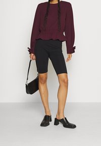 Burgunderfarbene Bluse mit langen Ärmeln und Peplum-Saum, kombiniert mit schwarzen Radlerhosen. Die Accessoires bestehen aus einer schwarzen Tasche mit Krokodil-Prägung und schwarzen Block-Absatz-Pantoletten.